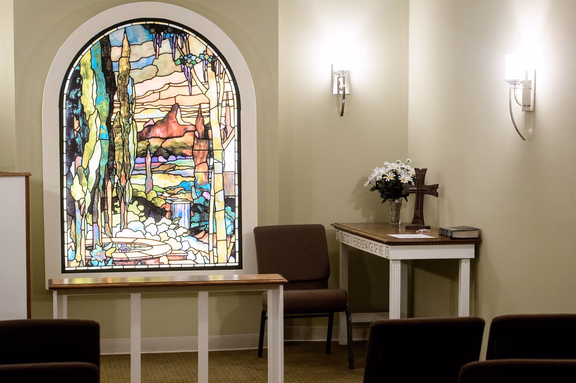Interior of a small chapel or prayer room with a large stained glass window depicting a colorful nature scene, a wooden bench, a brown chair, a table with a vase of white flowers, a wooden cross, and a book. The walls are light green with two modern wall sconces providing light.