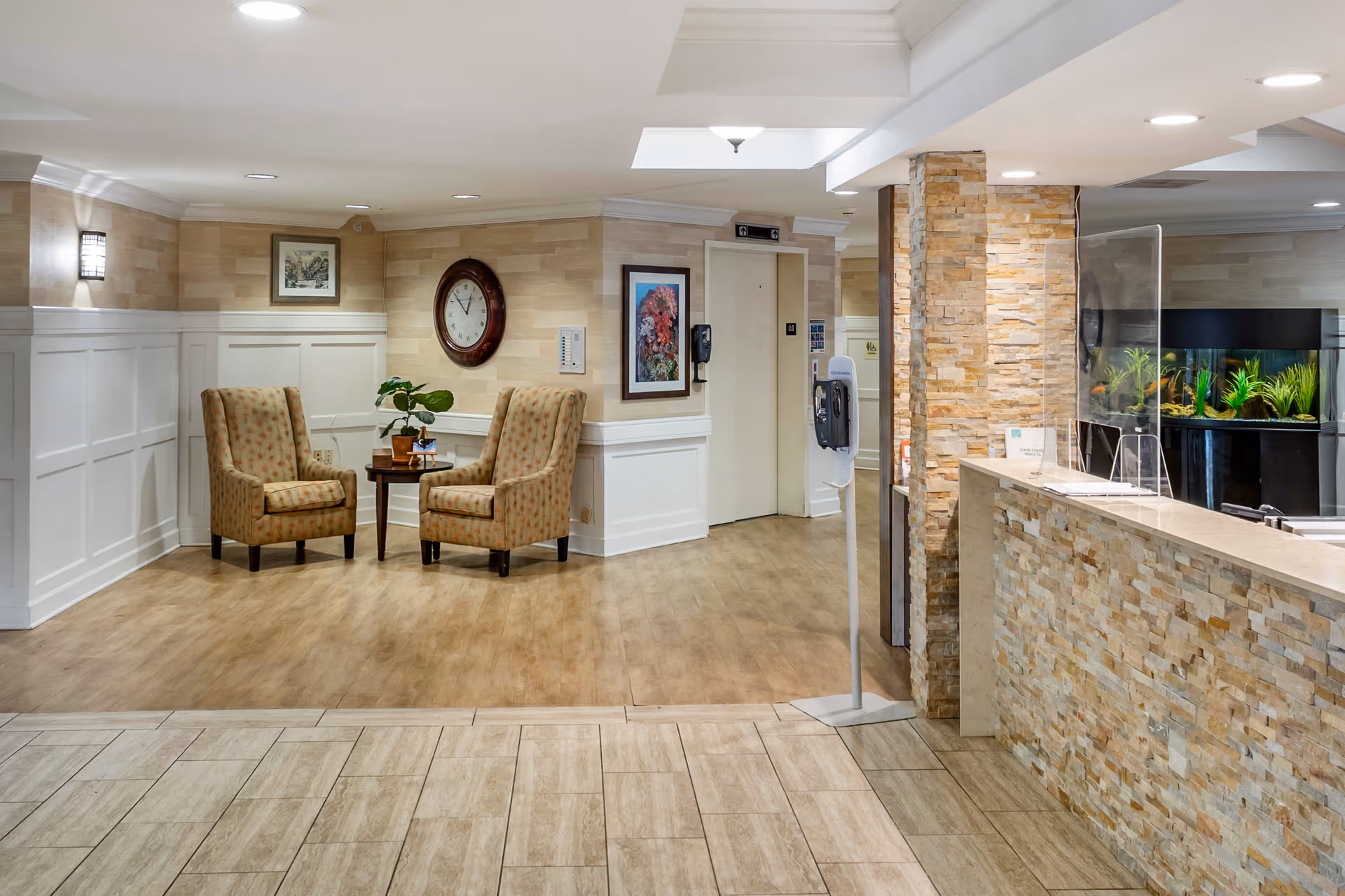 A well-lit interior lobby area featuring two patterned armchairs with a small round table between them, a wall clock, framed artwork, an elevator door, a hand sanitizer dispenser, and a reception desk with a stone facade and a clear protective screen. There is also a large fish tank behind the reception desk.