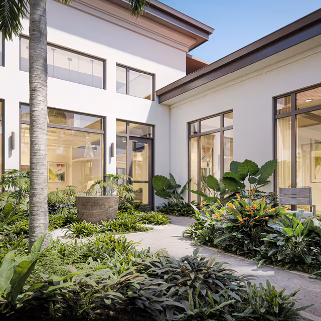 Sunlit courtyard with lush tropical plants, a paved walkway and large windows opening into a modern building.