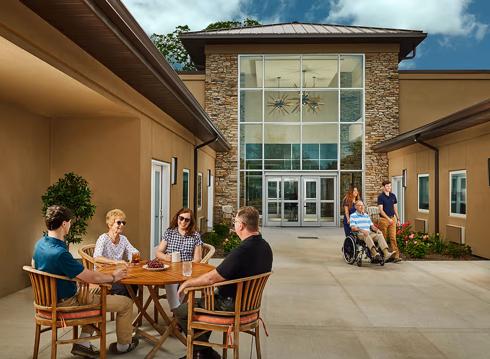 A group of people enjoying time outside a nursing home. Four individuals are seated around a wooden table with drinks and grapes, while two others are walking, one pushing a man in a wheelchair. The building has a modern design with large glass windows and stone accents under a partly cloudy sky.