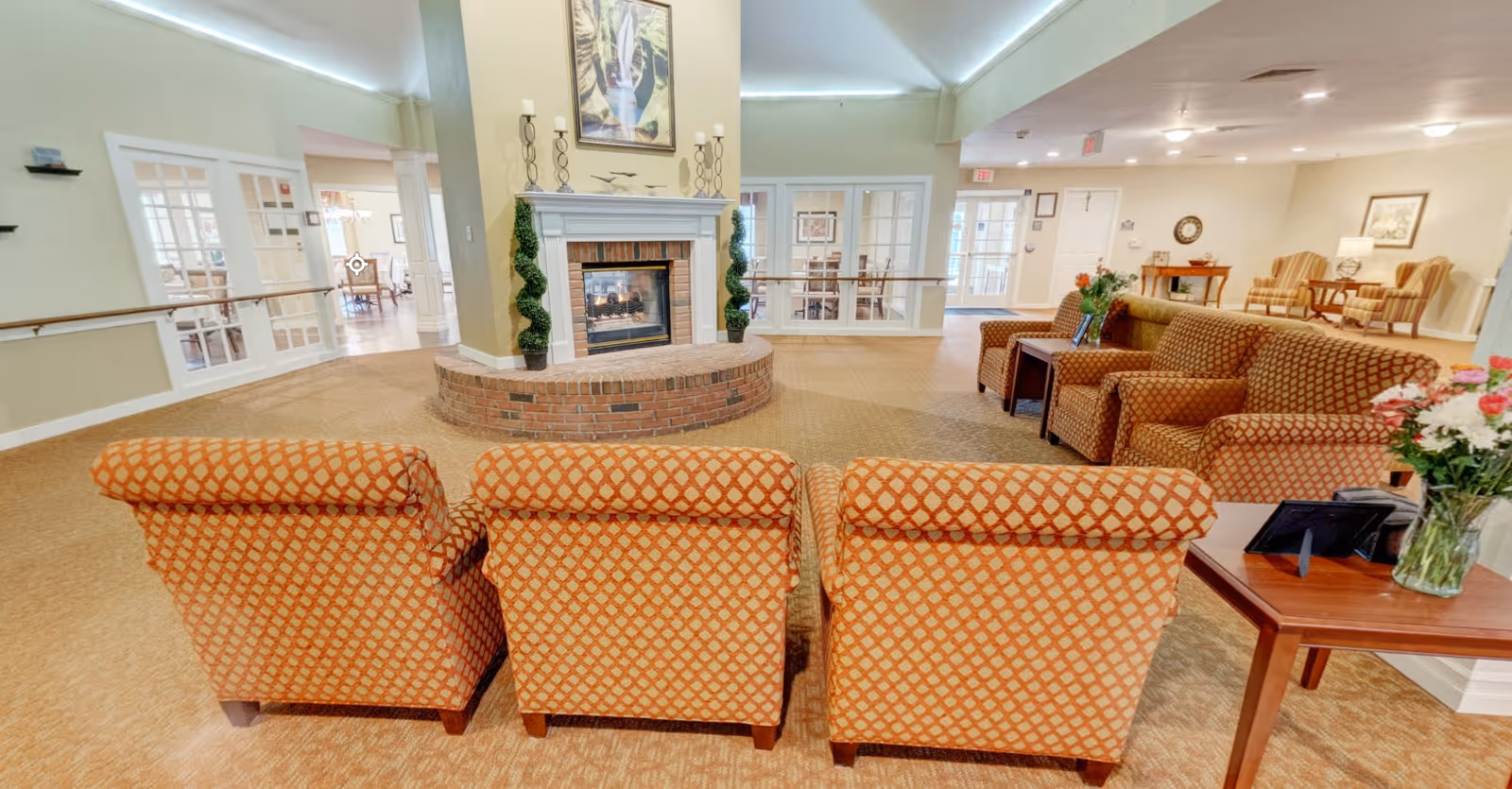 A spacious communal lounge with patterned armchairs arranged around a brick fireplace and additional seating and tables.