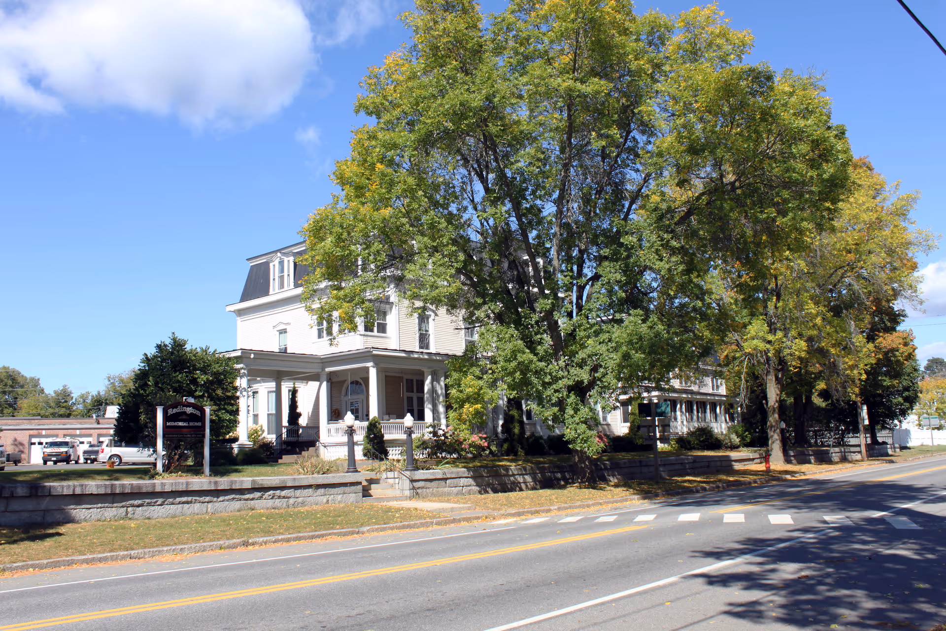 Large historic white building with a wraparound porch and tall trees set behind a low stone wall along a street.