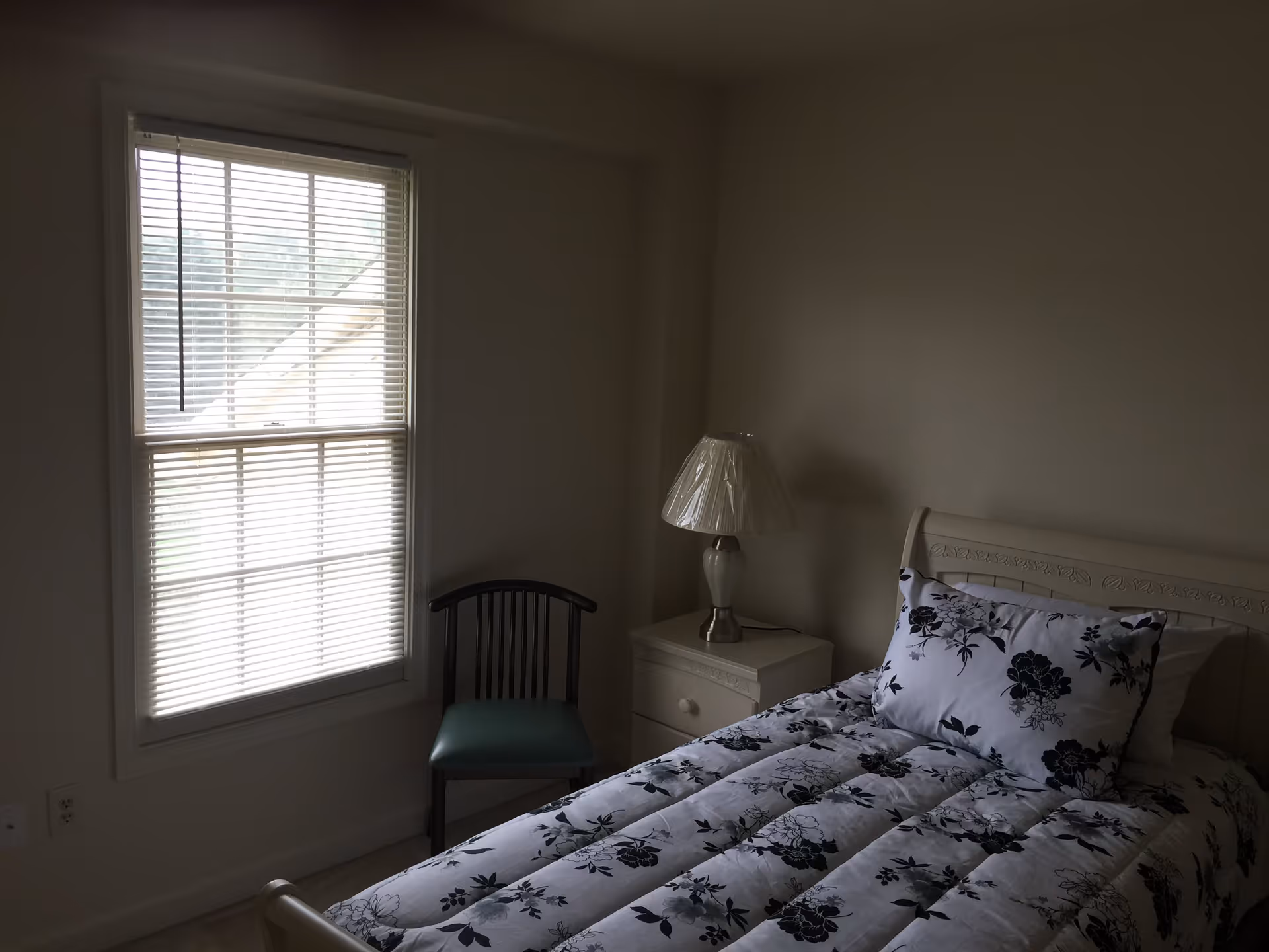 A small bedroom with a single bed covered in a white comforter with black floral patterns. There is a matching pillow on the bed. Next to the bed is a white nightstand with a lamp that has a white shade covered in plastic. A wooden chair with a green seat cushion is placed near a window with white blinds partially closed.