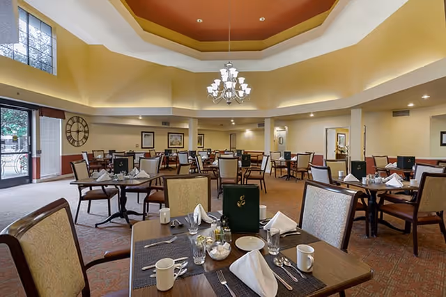 A spacious dining room with multiple tables set for meals, featuring chairs with cushioned seats and backs. The tables have place settings including napkins, cups, glasses, and silverware. The room has a high ceiling with a decorative recessed design and a chandelier hanging in the center. Large windows and glass doors allow natural light to enter the room. The walls are painted in warm tones, and there is a large clock on one wall.
