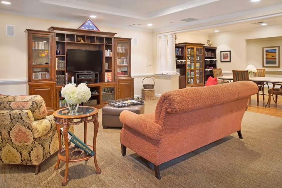 A cozy living room area in a senior living facility featuring a patterned armchair, a red upholstered sofa, a wooden side table with a vase of white flowers, and a large wooden entertainment center with a TV and shelves filled with books and decorative items. In the background, there is a dining area with a table and chairs, framed artwork on the walls, and a cabinet with glass doors displaying more items.