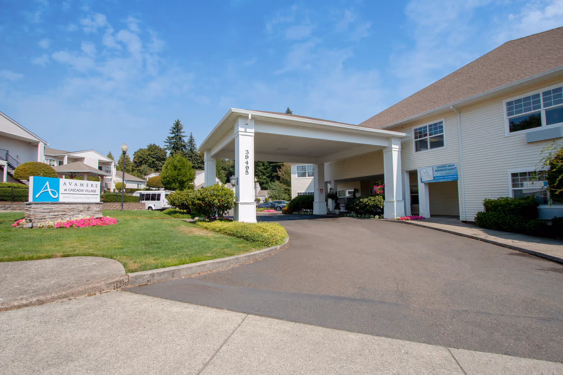 Exterior view of Cascadia Village Senior Living by Cogir showing the entrance driveway with a covered drop-off area, a sign for Avamere at Cascadia Village on a stone base surrounded by flowers, well-maintained green lawns, and residential buildings under a blue sky.