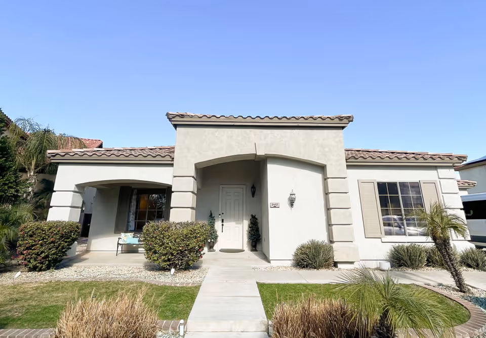 Single-story stucco house with an arched front porch, tiled roof, walkway and shrubs under a clear blue sky.