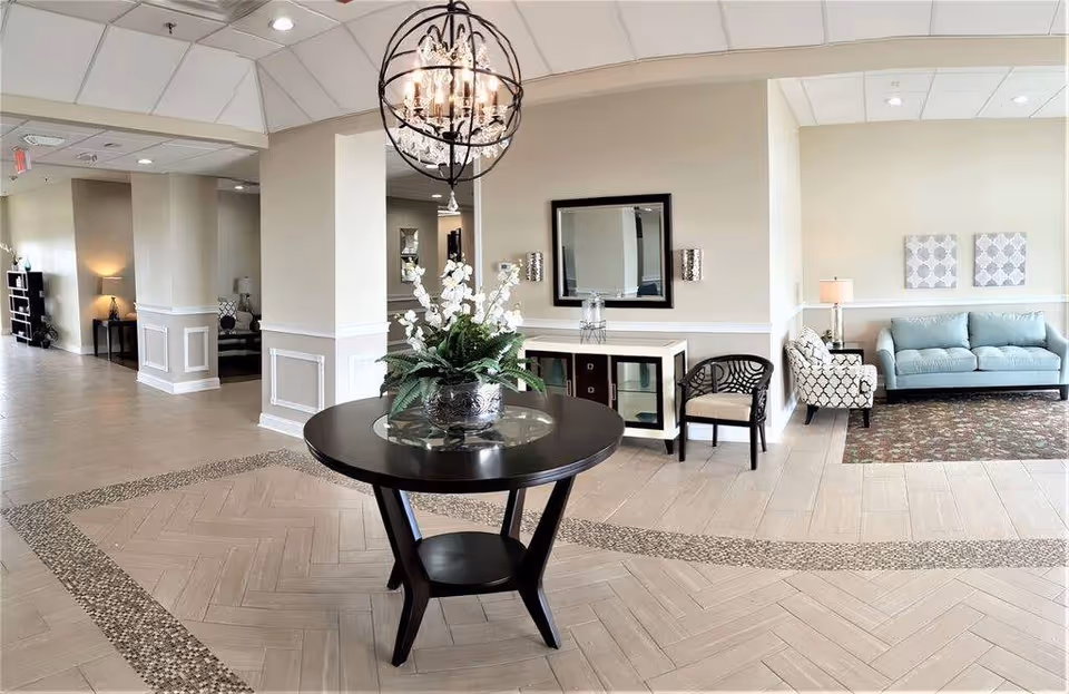 A spacious and well-lit interior common area with a round black table holding a floral arrangement in the center. The room features beige walls with white trim, a chandelier hanging from the ceiling, a console table with a mirror above it, and seating including a patterned armchair and a light blue sofa on a patterned rug.