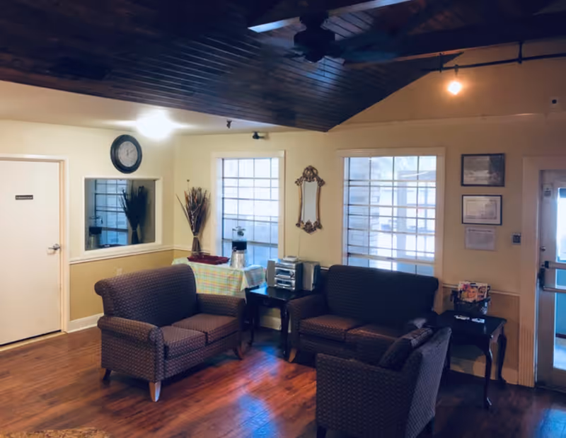 A cozy living room area with wooden floors and a dark wooden ceiling. The room features two patterned loveseats and a matching armchair arranged around a small table with a stereo system. There are two large windows letting in natural light, a decorative mirror on the wall, a clock above a small window, and a door labeled 'Restroom'. A table covered with a checkered cloth holds a coffee pot and cups.