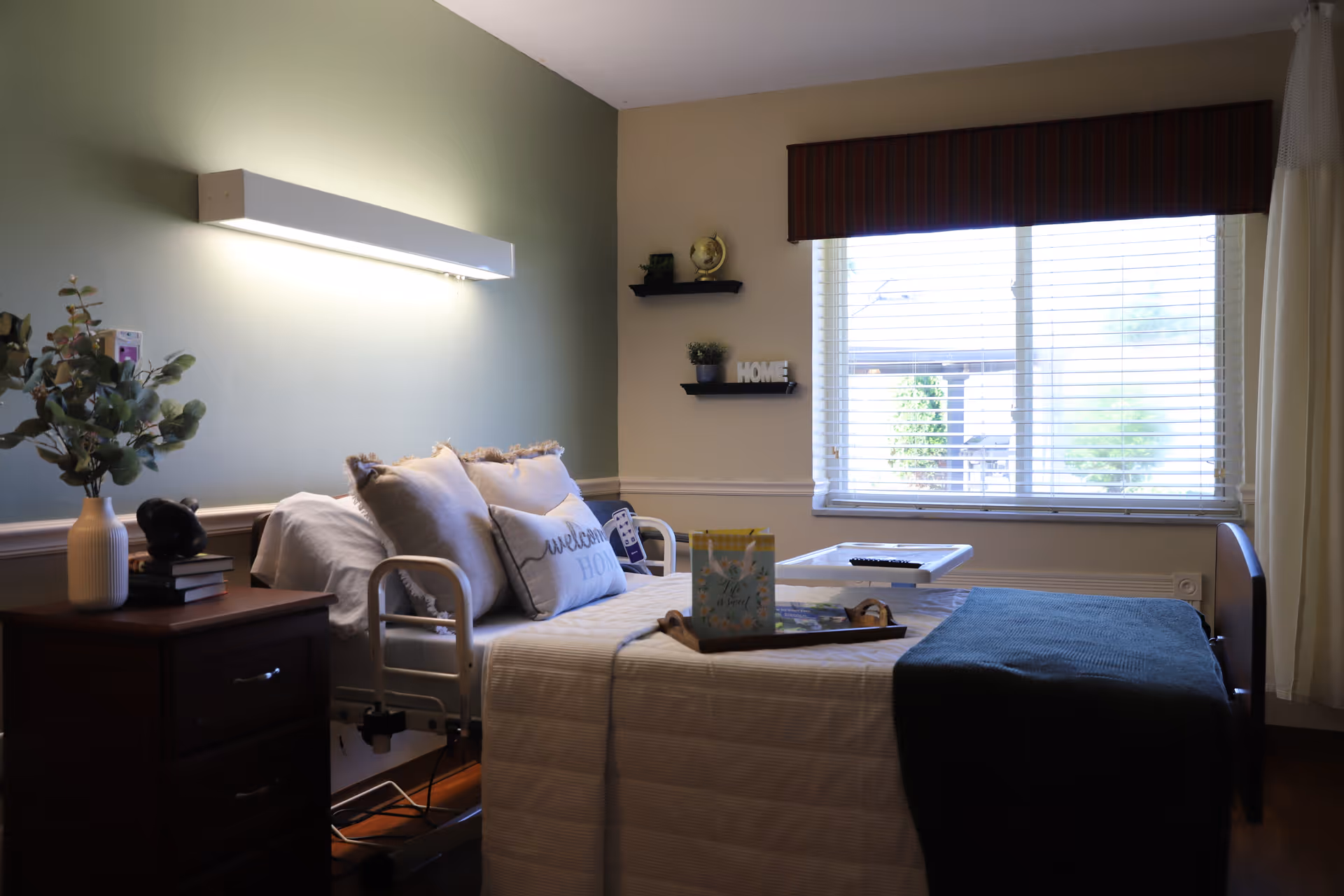 A cozy bedroom in a senior living facility with a hospital-style bed adorned with multiple pillows, including one that says 'welcome HOME'. There is a wooden nightstand with a vase of greenery and books, a wall-mounted light fixture above the bed, two small shelves on the wall with decorative items, and a large window with blinds letting in natural light.