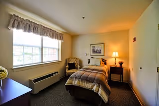 A cozy bedroom with a single bed covered in a plaid blanket, a wooden nightstand with a lamp, a cushioned armchair, a framed picture on the wall, a large window with a valance, and a dresser with a vase of flowers.