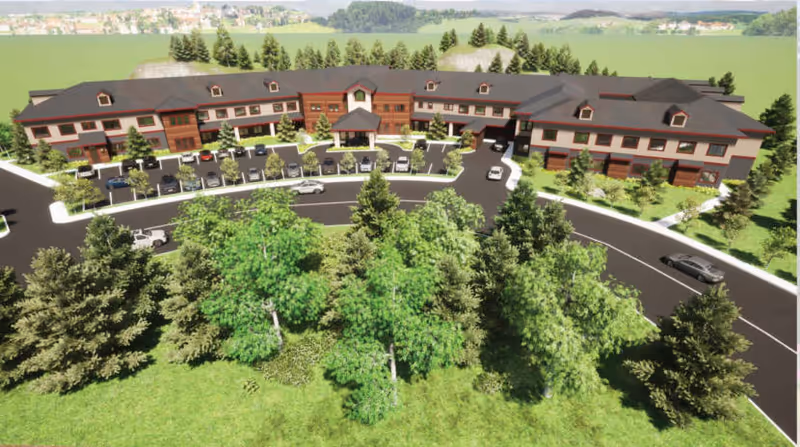 Aerial view of a two-story senior living building with a circular drive, parked cars, and trees in the foreground.