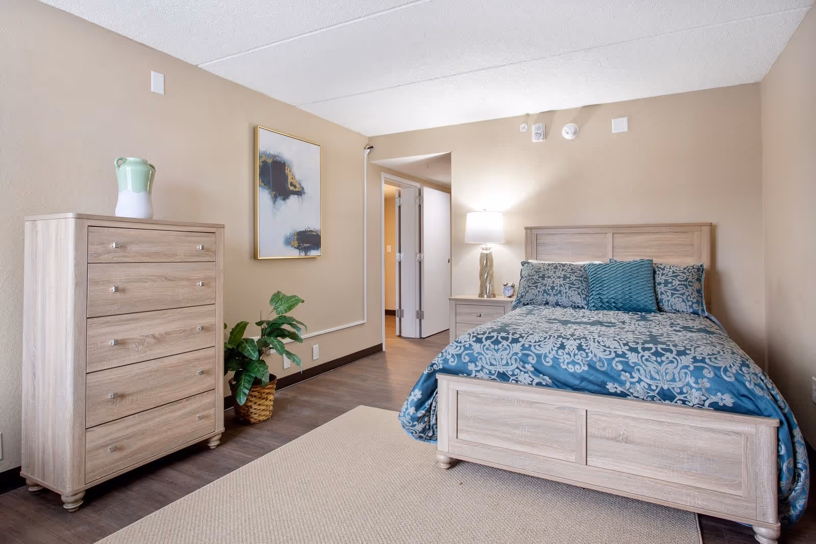A bedroom with a wooden bed frame and a blue patterned comforter with matching pillows. Next to the bed is a wooden nightstand with a lamp and a small clock. On the left side of the room is a wooden chest of drawers with a green and white vase on top. A potted plant is placed on the floor beside the chest. The walls are beige, and there is a framed abstract painting hanging above the plant. The floor is a combination of wood and a beige rug. An open doorway leads to another room.