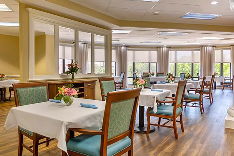 A bright and spacious dining room with multiple tables covered in white tablecloths, each set with a small vase of flowers and blue napkins. The room features large windows with beige curtains, allowing natural light to fill the space. Wooden chairs with teal patterned cushions surround the tables, and the floor is a warm-toned wood.