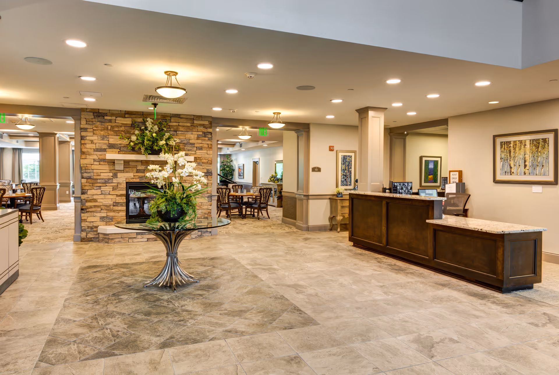 Spacious and well-lit senior living facility lobby area with a stone fireplace in the center, a round glass table with a floral arrangement, a wooden reception desk on the right, and dining tables with chairs visible in the background.