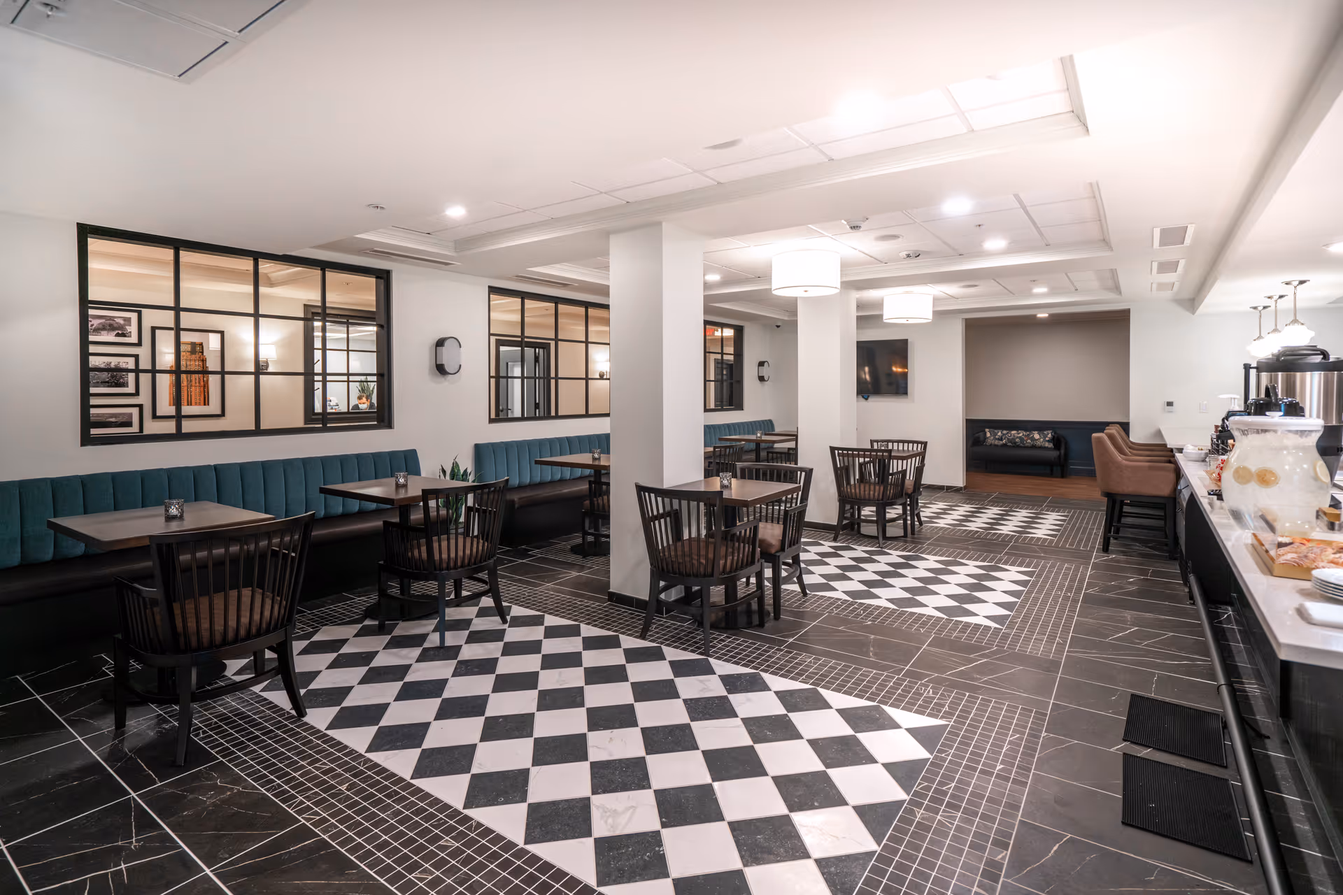 A modern dining area with black and white checkered floor tiles, several small square tables with dark wooden chairs, and teal upholstered bench seating along the wall. The room has white walls with large windows looking into another room, ceiling lights, and a counter on the right side with refreshments and plates.