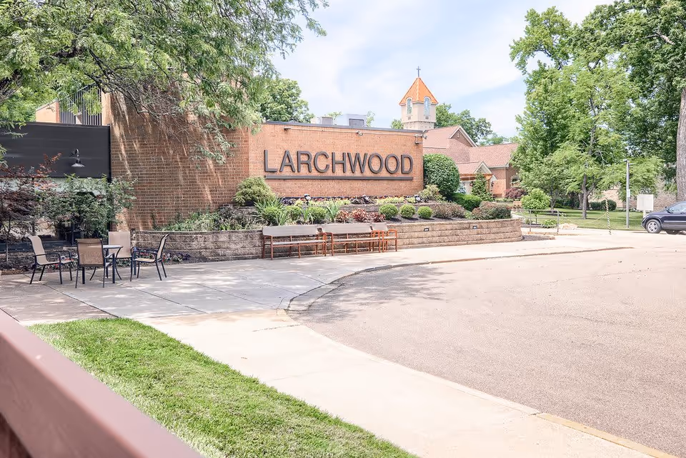 Outdoor view of a senior living facility named Larchwood with a brick wall sign, surrounded by landscaped bushes and trees. There are several chairs and tables on a paved area near the sign, and a building with a tower and cross is visible in the background under a partly cloudy sky.
