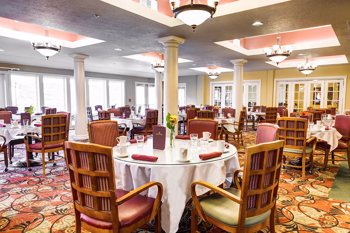 A spacious dining room with multiple round tables covered with white tablecloths, each set with cups, glasses, and folded red napkins. The room features wooden chairs with red and striped upholstery, decorative columns, patterned carpet, and ceiling lights with recessed lighting accents.