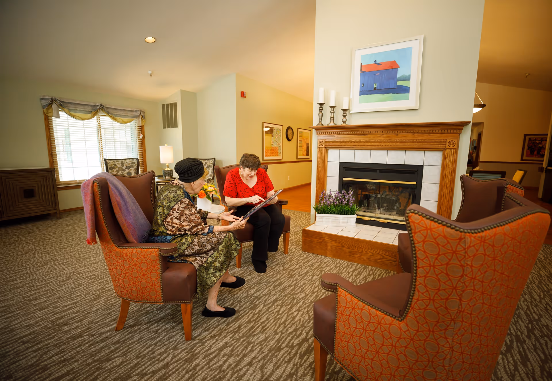 Two elderly women sitting in armchairs in a cozy living room area with a fireplace, looking at a photo album together. The room has patterned carpet, a window with curtains, framed artwork on the walls, and decorative candles on the fireplace mantel.