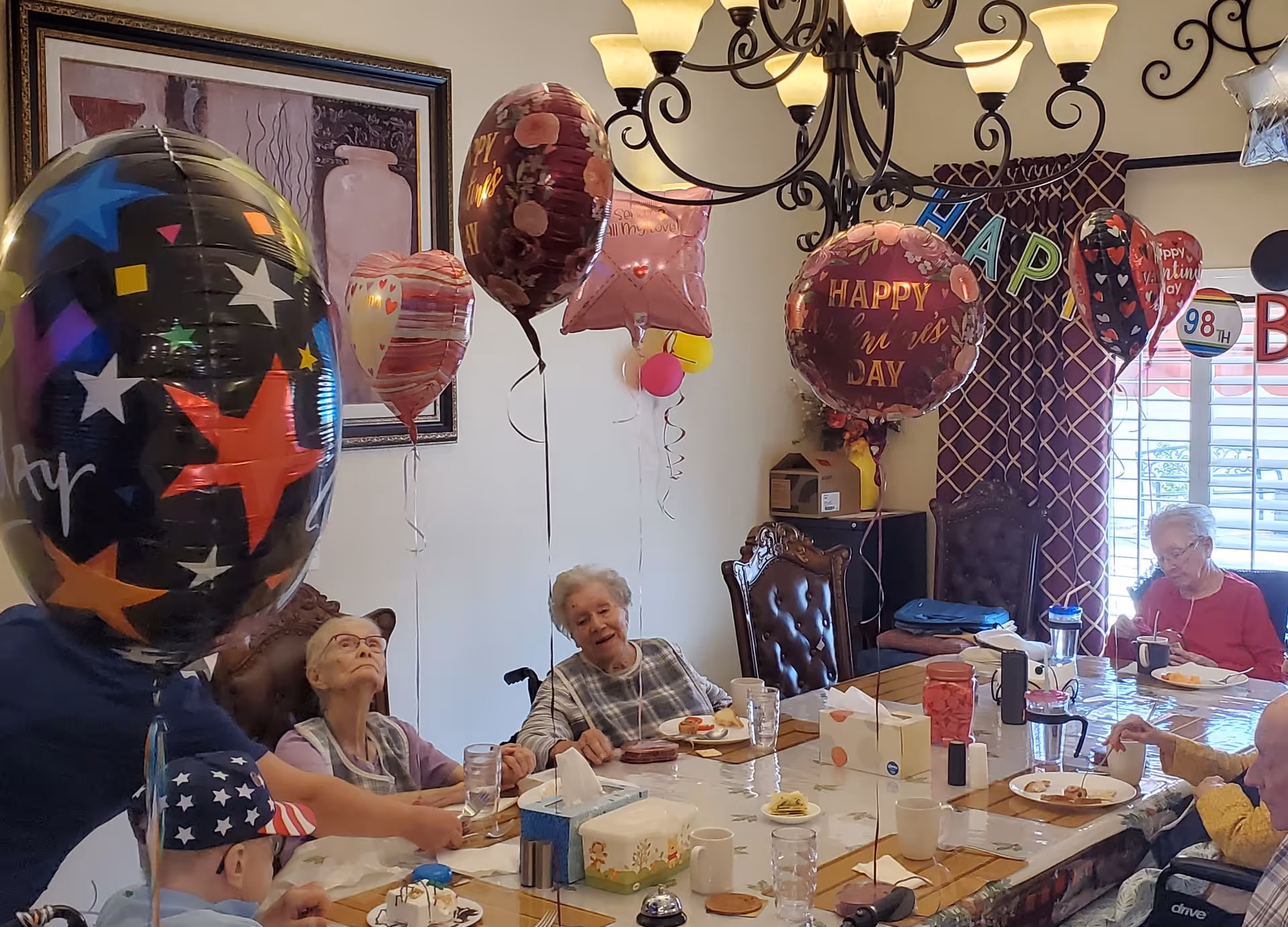A group of elderly people sitting around a dining table decorated with balloons for a celebration. The table has plates with food, cups, and tissue boxes. The room has a chandelier, framed artwork on the wall, and a window with patterned curtains. Some balloons have messages like 'Happy Nurses Day' and 'Happy Valentine's Day'.