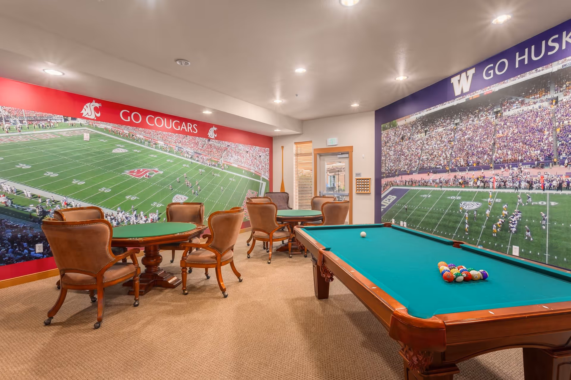 A recreational room with a pool table and two round card tables surrounded by brown leather chairs. The walls feature large murals of football stadiums with one side showing 'GO COUGARS' and the other side showing 'GO HUSKIES'. The room has beige carpet and recessed ceiling lights.