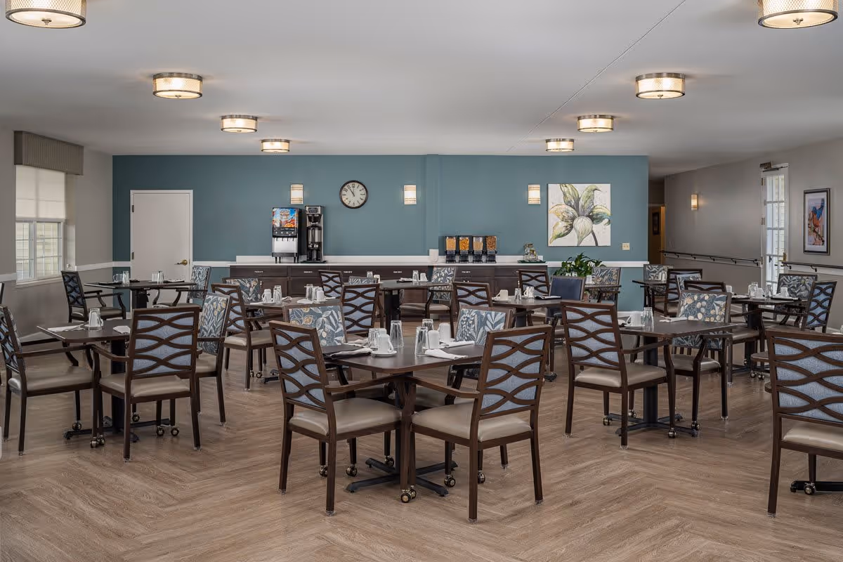 A spacious dining room with multiple dark wood tables and chairs with patterned cushions. The room has a light blue accent wall with a clock, a coffee and beverage station, and a large floral painting. The floor is wood with a herringbone pattern, and the ceiling has several round light fixtures. Windows with blinds allow natural light into the room.