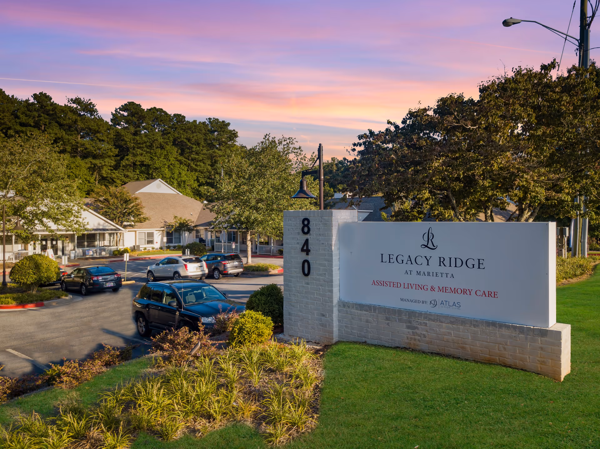 View of the entrance sign for Legacy Ridge at Marietta, an assisted living and memory care facility, with a parking lot and buildings surrounded by trees under a colorful sunset sky.