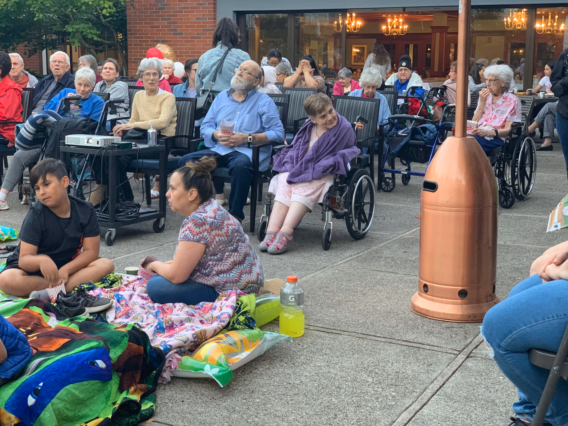 A group of elderly people, some in wheelchairs, and a few younger individuals are gathered outdoors on a concrete patio. Some people are seated on chairs while others sit on blankets on the ground. There is a copper-colored outdoor heater in the foreground and a brick building with large windows and chandeliers visible inside in the background.