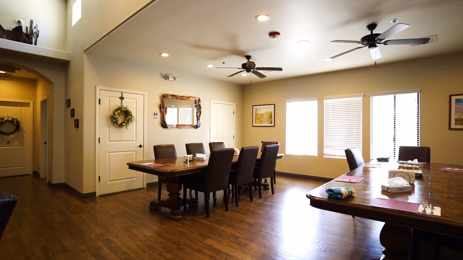 A dining room with two large wooden tables surrounded by brown leather chairs. The room has wooden flooring, beige walls, ceiling fans, recessed lighting, and three windows with blinds. There are decorative wreaths on two doors and framed artwork on the walls.