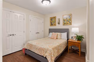 A bedroom with a double bed featuring a patterned bedspread and several pillows. There are two framed artworks hanging above the bed. To the right of the bed is a wooden nightstand with a white lamp, a small plant, and framed photos. The room has light-colored walls, a ceiling light fixture, and two closed white double-door closets.