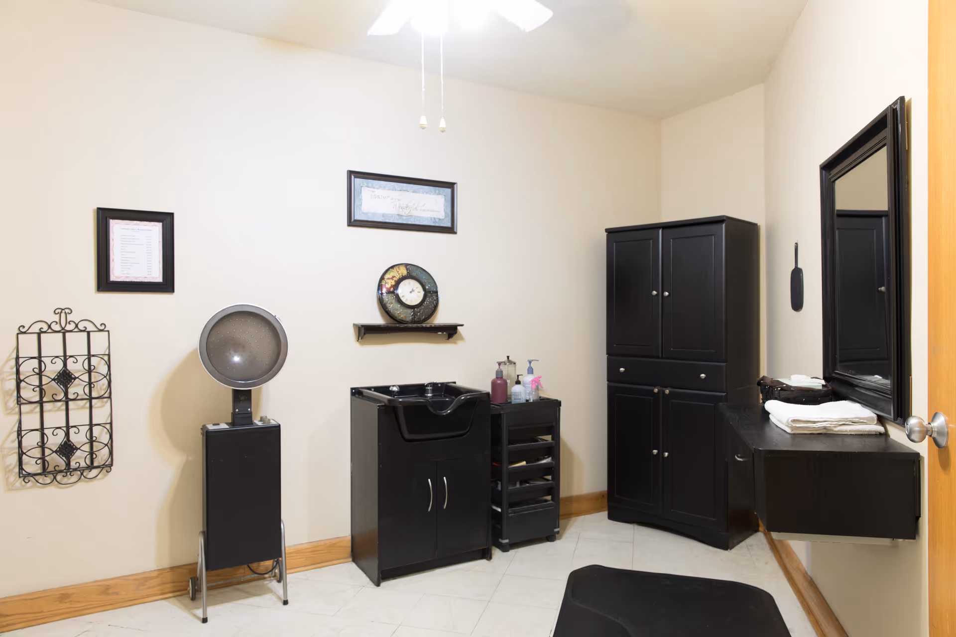 Interior of a small salon or hair care room with a black hair dryer chair, a black sink cabinet, a black storage cabinet, a wall-mounted mirror, and various hair care products on a small rolling cart. The walls are light-colored with wooden baseboards and there are framed decorations on the wall.