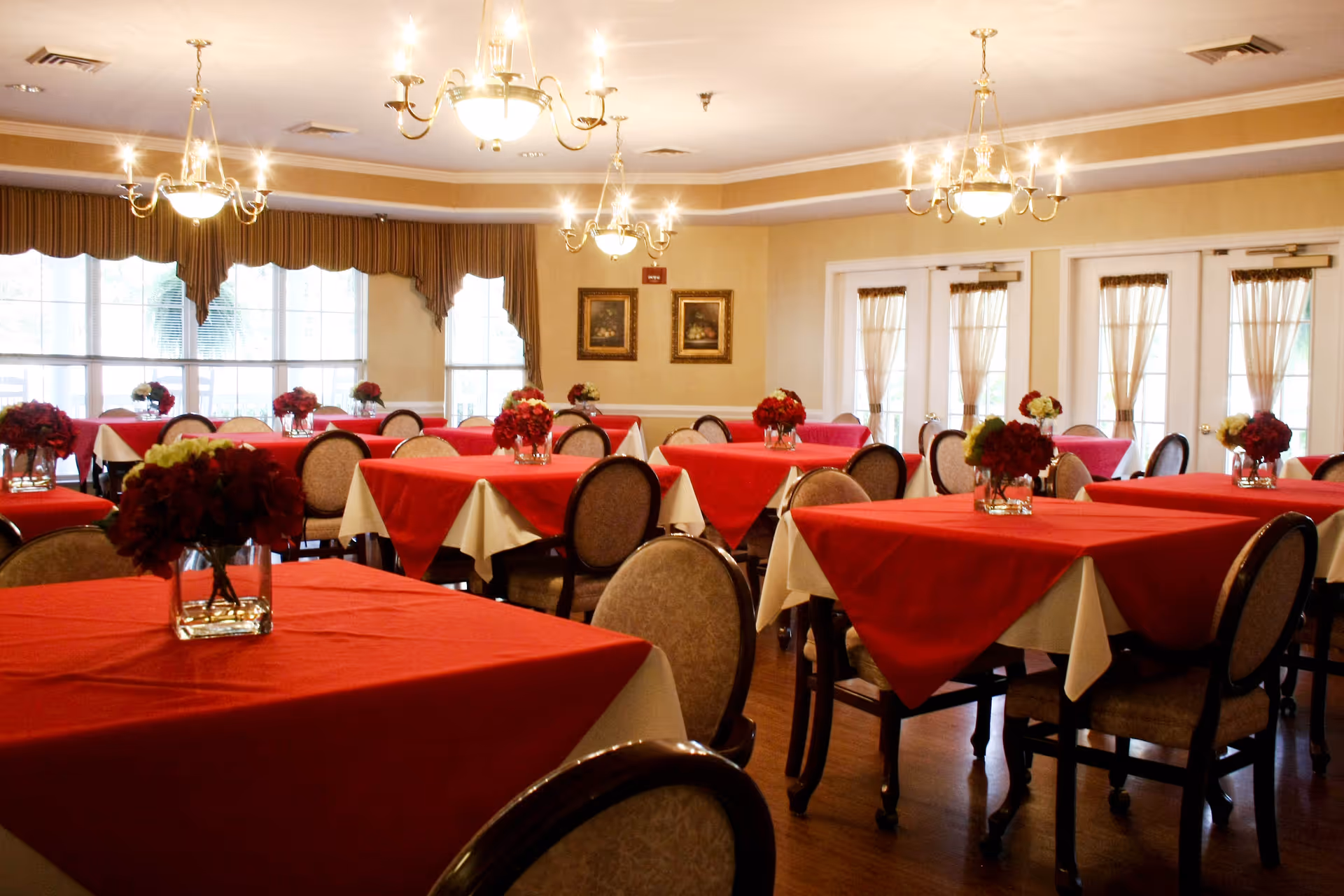 A dining room with multiple tables covered in white tablecloths and red overlays, each adorned with a vase of red and white flowers. The room has large windows with brown curtains, chandeliers hanging from the ceiling, and framed artwork on the walls.