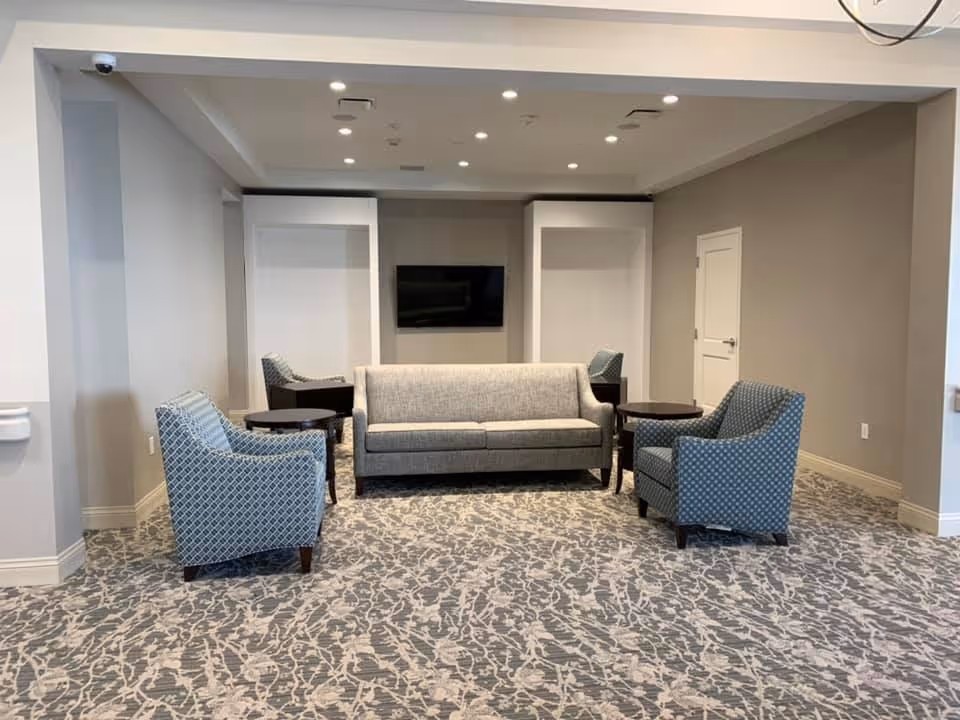 A neutral-toned common sitting area with a gray sofa, patterned armchairs, side tables, and a wall-mounted TV.