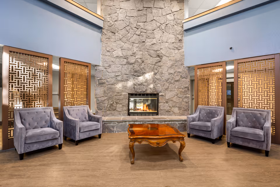 A cozy seating area in a senior living community featuring four gray upholstered armchairs arranged around a polished wooden coffee table. Behind the seating is a large stone fireplace with a fire burning, flanked by decorative wooden lattice panels. The walls are painted light blue, and the floor has a wood-like finish.
