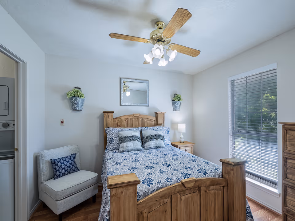 Well-lit bedroom with a wooden bed dressed in blue floral bedding, an armchair, nightstand and window with blinds.