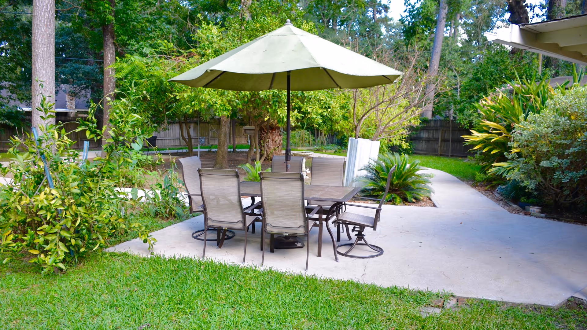 Outdoor patio area with a rectangular table and six chairs under a large beige umbrella, surrounded by green grass, trees, and various plants with a concrete pathway curving around the seating area.