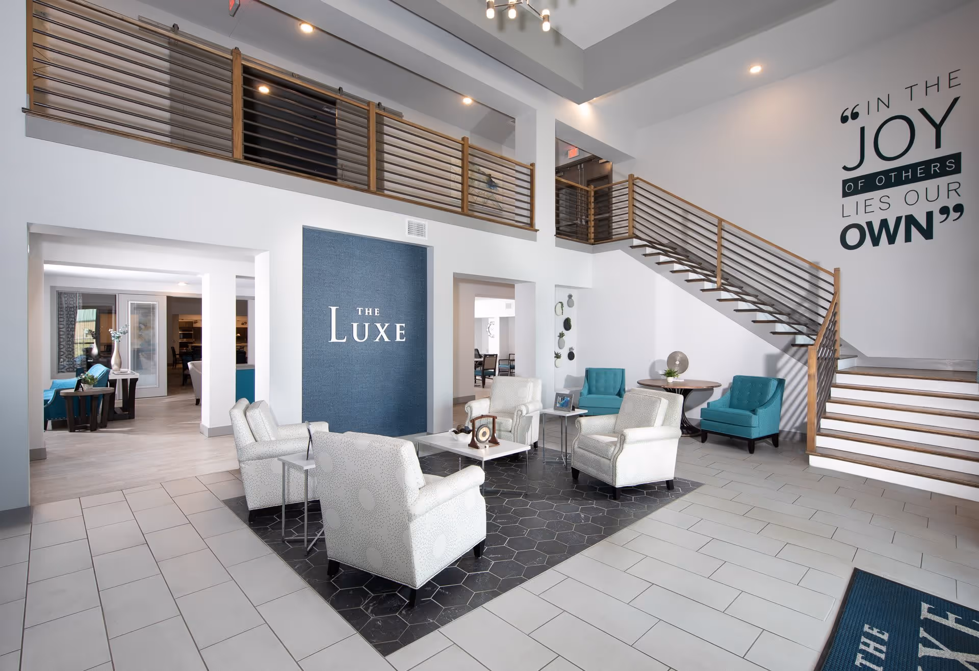 Bright modern lounge lobby with armchairs around a central table, teal accent chairs, a staircase, and a wall sign reading "The Luxe".