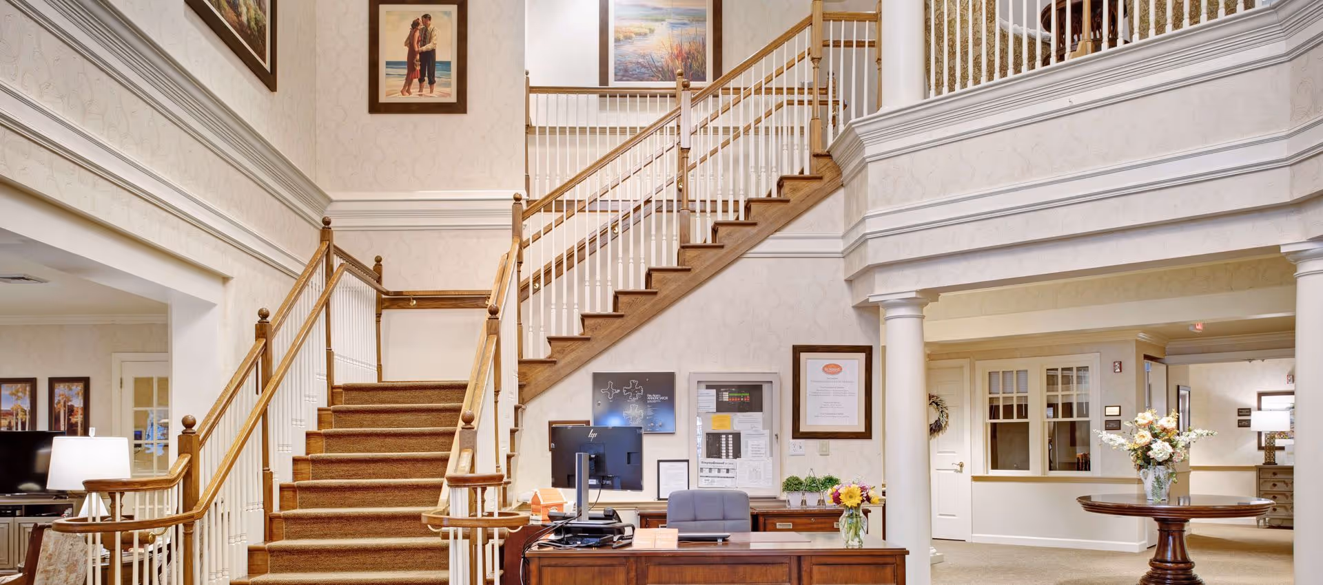 Interior view of a senior living facility lobby with a wooden staircase featuring white balusters and brown carpeted steps. A reception desk with a computer and flowers is positioned in front of the stairs. The walls are decorated with framed artwork and certificates. To the right, there is a round wooden table with a vase of flowers, and the area is well-lit with warm lighting.