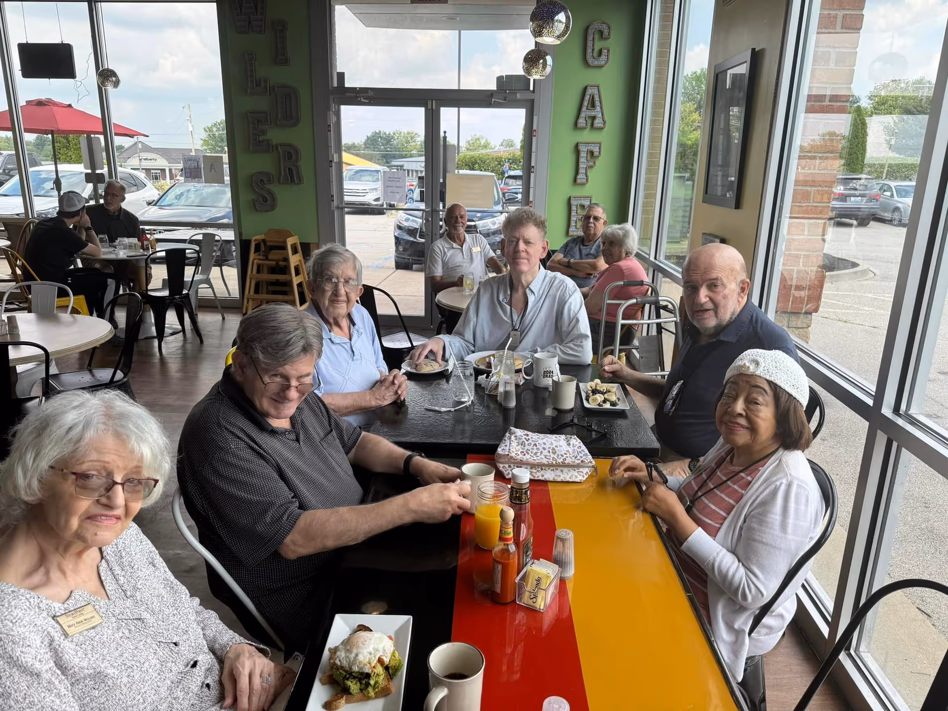 A group of elderly people sitting around a table in a bright café with large windows. They are enjoying food and drinks, smiling and engaging with each other. The café has a casual atmosphere with other patrons visible in the background and a view of the parking lot outside.