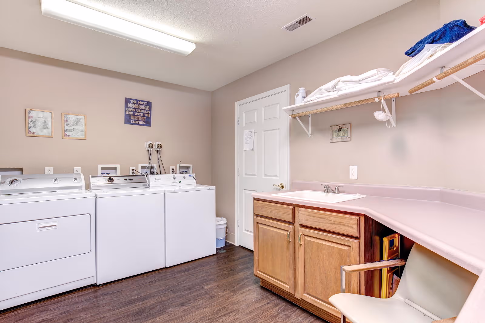 Laundry room with a row of washers and a dryer, a sink set in a countertop with wooden cabinets, and shelving holding linens.