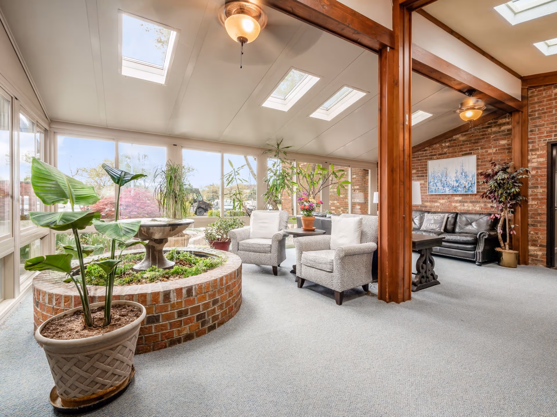 A bright and spacious indoor lounge area with large windows and skylights allowing natural light to fill the space. The room features a circular brick planter with green plants and a small fountain in the center. There are two upholstered armchairs with a small table between them, a black leather sofa against a brick wall, and various potted plants. Wooden beams and a ceiling fan add to the cozy atmosphere.