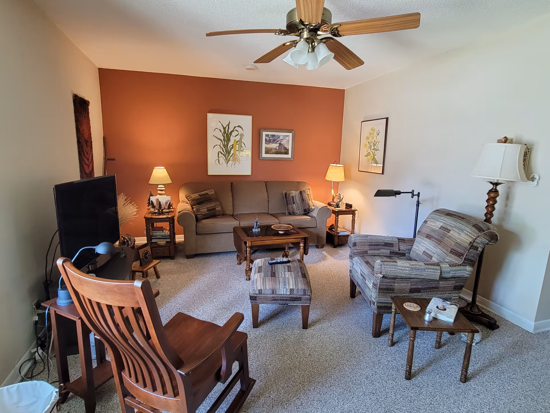 Cozy living room with a sofa against a rust-colored accent wall, patterned armchair and ottoman, wooden chairs, coffee tables, lamps, TV, and ceiling fan.