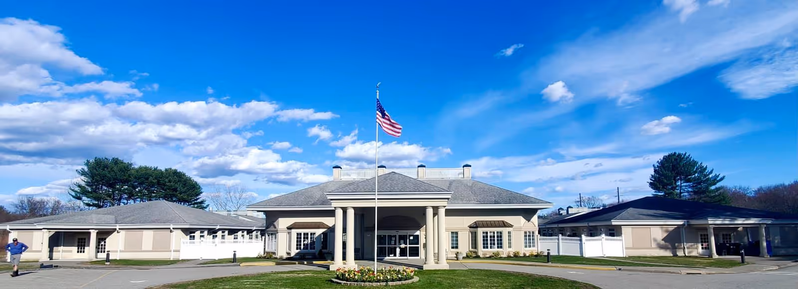 Front exterior view of Greentree Manor Nursing and Rehabilitation building with a flagpole flying the American flag in front, surrounded by a circular flower bed and driveway under a blue sky with scattered clouds.