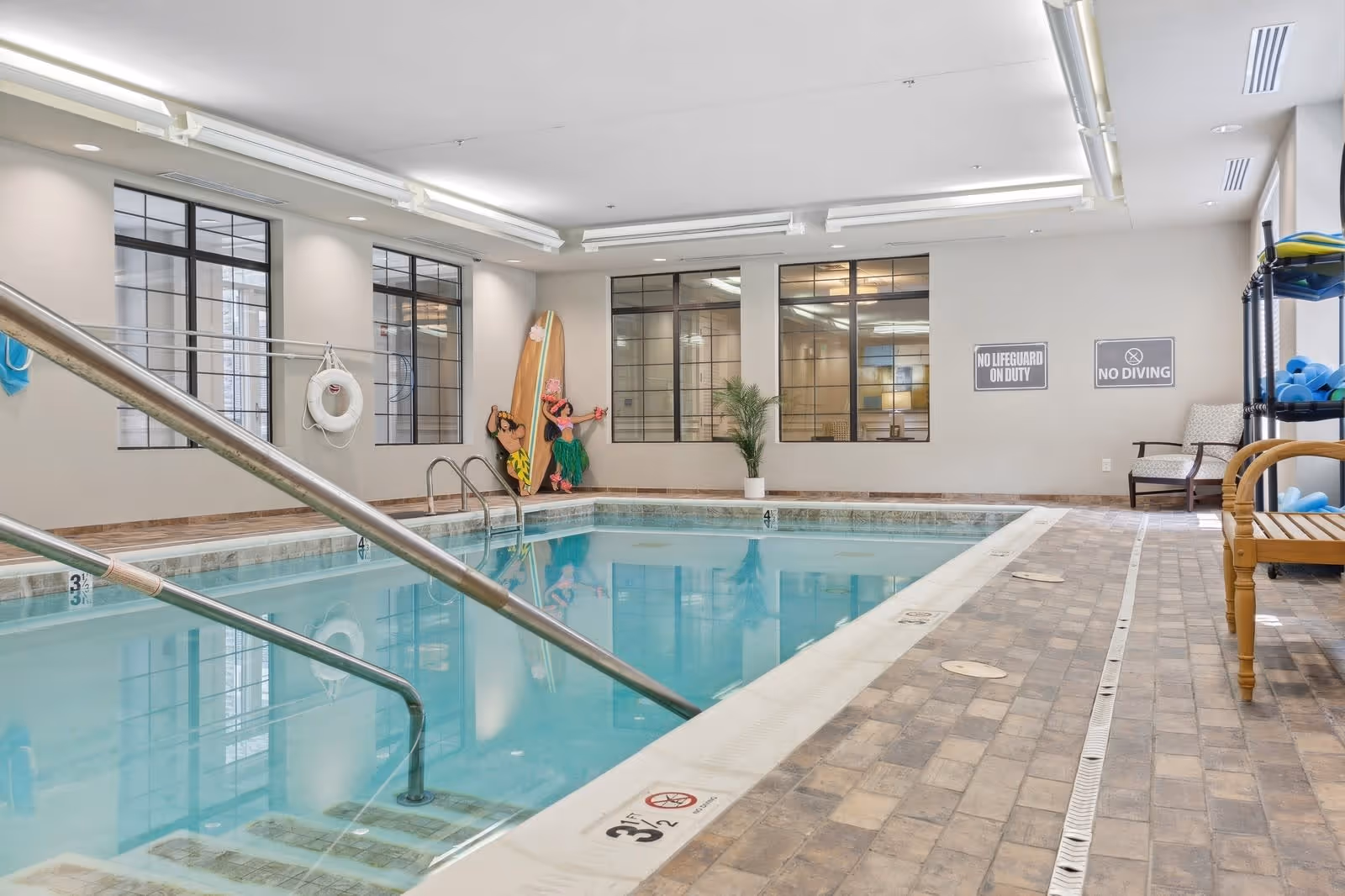 Indoor swimming pool area with handrails leading into the water, tiled floor, large windows, safety signs on the wall, a life preserver, and pool equipment on shelves. There is also a decorative surfboard and tropical-themed wall art.