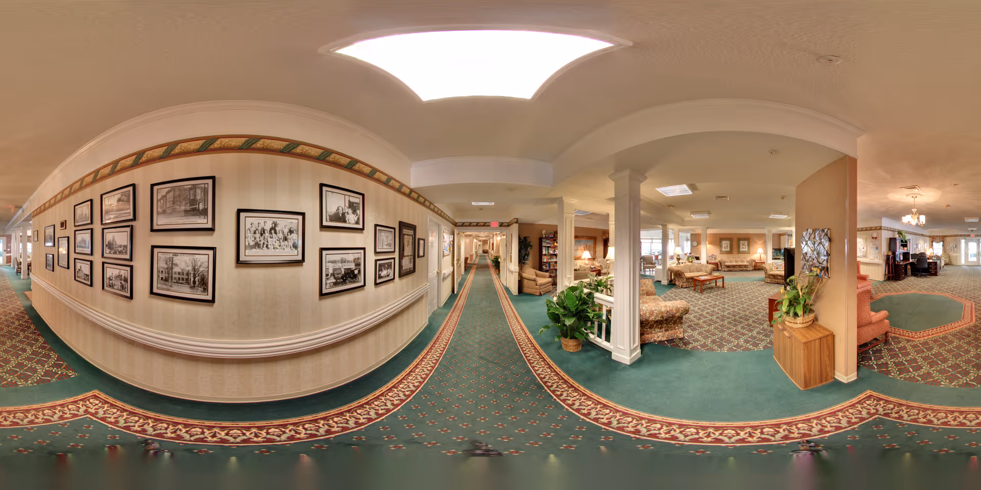 A wide panoramic view of an interior hallway and lounge area in a senior living facility. The hallway has green carpet with a red and gold patterned border, cream-colored walls adorned with framed black and white photographs. The lounge area features comfortable seating including armchairs and sofas, plants, and soft lighting. The space has a warm and inviting atmosphere with decorative ceiling lights and columns.