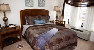 A cozy bedroom with a wooden headboard bed dressed in a patterned brown and blue comforter, pillows, and a folded purple throw blanket. There are two matching bedside tables with lamps, a pair of slippers on the carpeted floor, and a window with sheer white curtains and a dark valance letting in natural light.