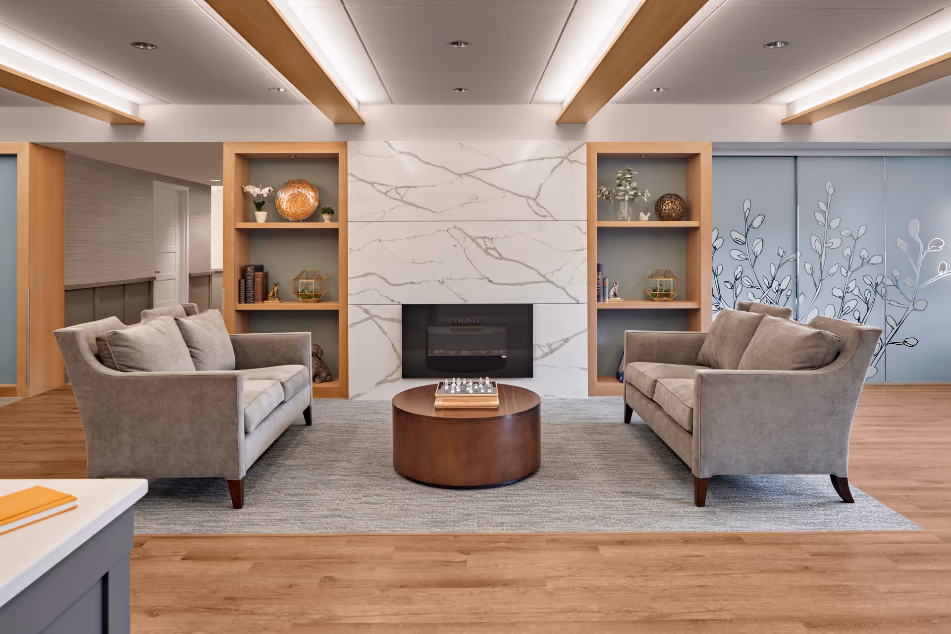 A modern living room with two gray sofas facing each other across a round wooden coffee table with a chessboard on it. The room features a marble fireplace centered between two wooden shelves decorated with books, plants, and decorative items. The floor is wooden with a large gray area rug, and the ceiling has recessed lighting with wooden beams. A wall with a floral design is visible in the background.