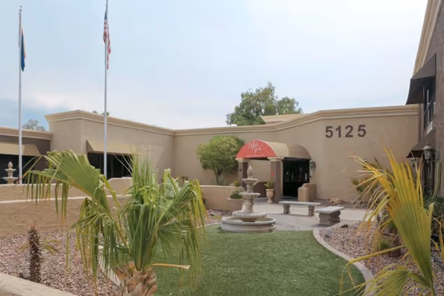 Exterior view of Bella Vita Health & Rehabilitation Center showing the building entrance with the number 5125 above it, a red awning, a small fountain, benches, and landscaping with palm plants and gravel.