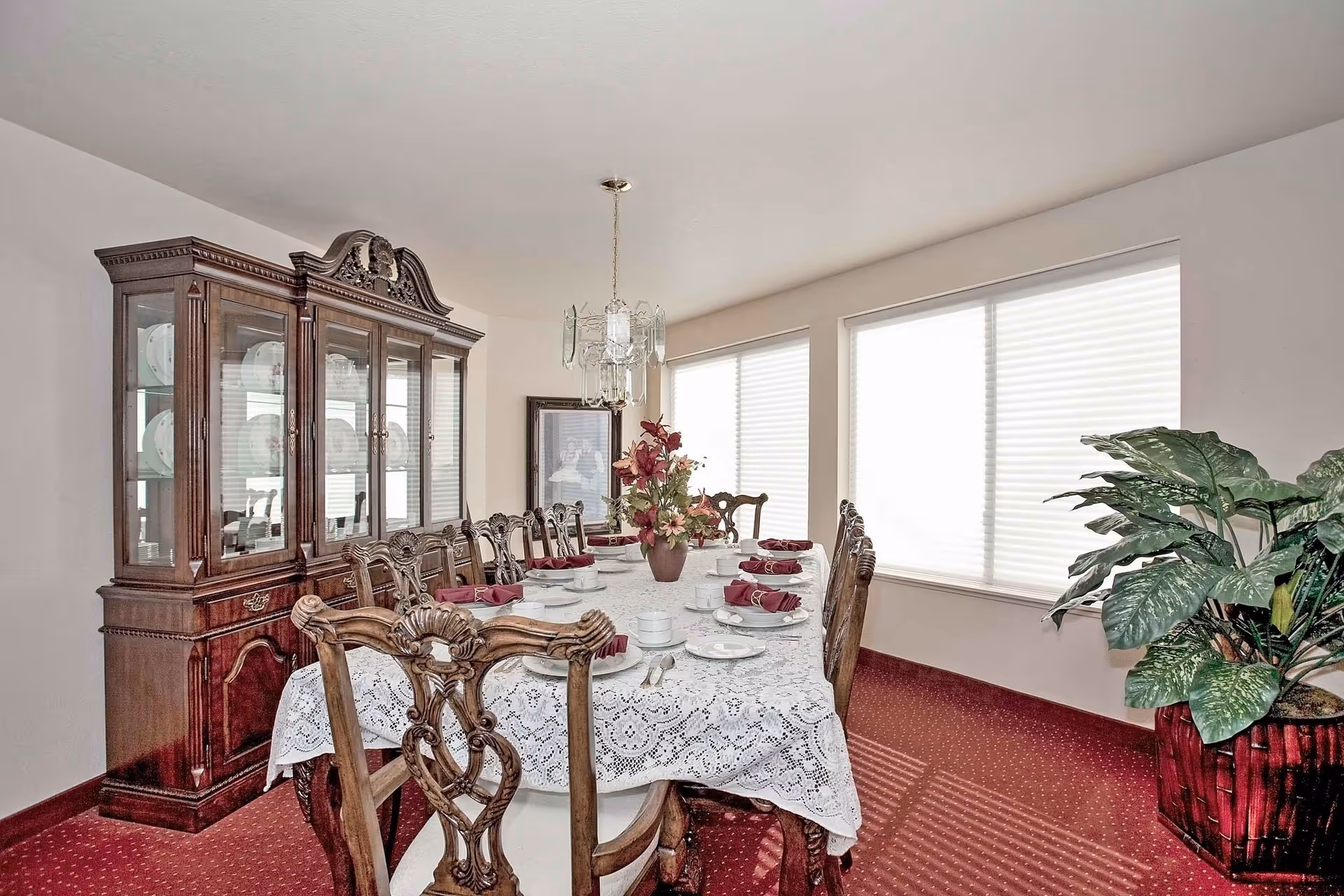A formal dining room with a long wooden dining table covered with a white lace tablecloth, set with white plates, cups, and burgundy napkins. The room features ornate wooden chairs, a large wooden china cabinet displaying plates, two large windows with blinds, a chandelier hanging above the table, a framed picture on the wall, and a large potted plant in the corner on a red carpeted floor.
