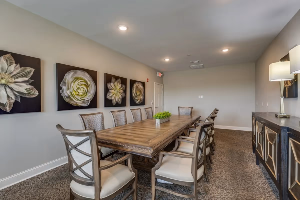 A long wooden dining table with cushioned chairs arranged around it in a well-lit room. The walls are decorated with large floral artwork, and a sideboard with a lamp is visible on the right side of the room.